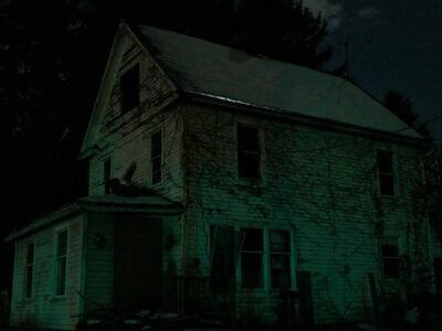 Casa de madeira abandonada à noite, iluminada por uma luz esverdeada entre árvores escuras