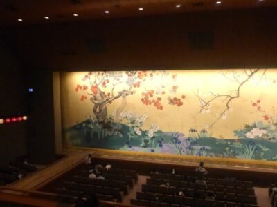 Plateia de teatro ocupada diante do pano de boca fechado, pintado com arvores e flores, antes do inicio do espetaculo.