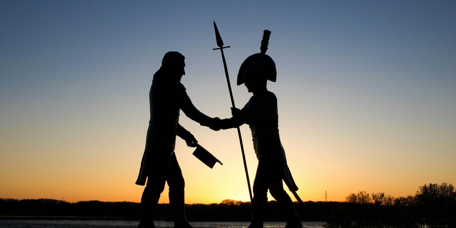 Silhueta de um monumento ao entardecer, com duas figuras apertando as mãos enquanto uma delas segura uma lança e a outra carrega um chapéu e uma arma.