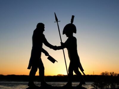Silhueta de um monumento ao entardecer, com duas figuras apertando as mãos enquanto uma delas segura uma lança e a outra carrega um chapéu e uma arma.