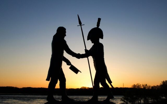 Silhueta de um monumento ao entardecer, com duas figuras apertando as mãos enquanto uma delas segura uma lança e a outra carrega um chapéu e uma arma.