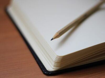 Caderno aberto em página em branco sobre mesa de madeira, com um lápis apoiado na folha da direita.