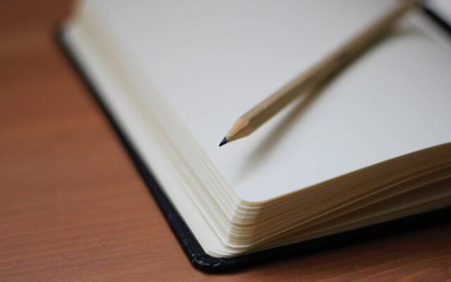 Caderno aberto em página em branco sobre mesa de madeira, com um lápis apoiado na folha da direita.