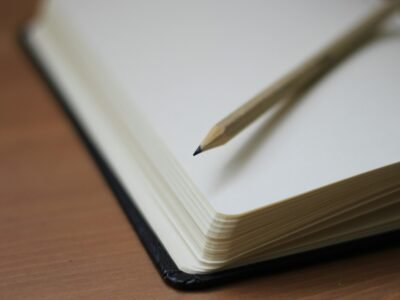 Caderno de capa preta aberto em página em branco sobre mesa de madeira, com um lápis apontado repousado sobre o papel